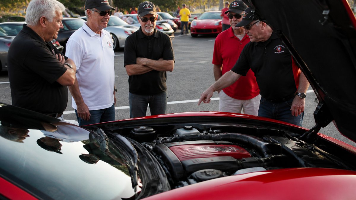 Corvette owners gather to network with enthusiasts while discussing engine details at a sunny local car meetup.