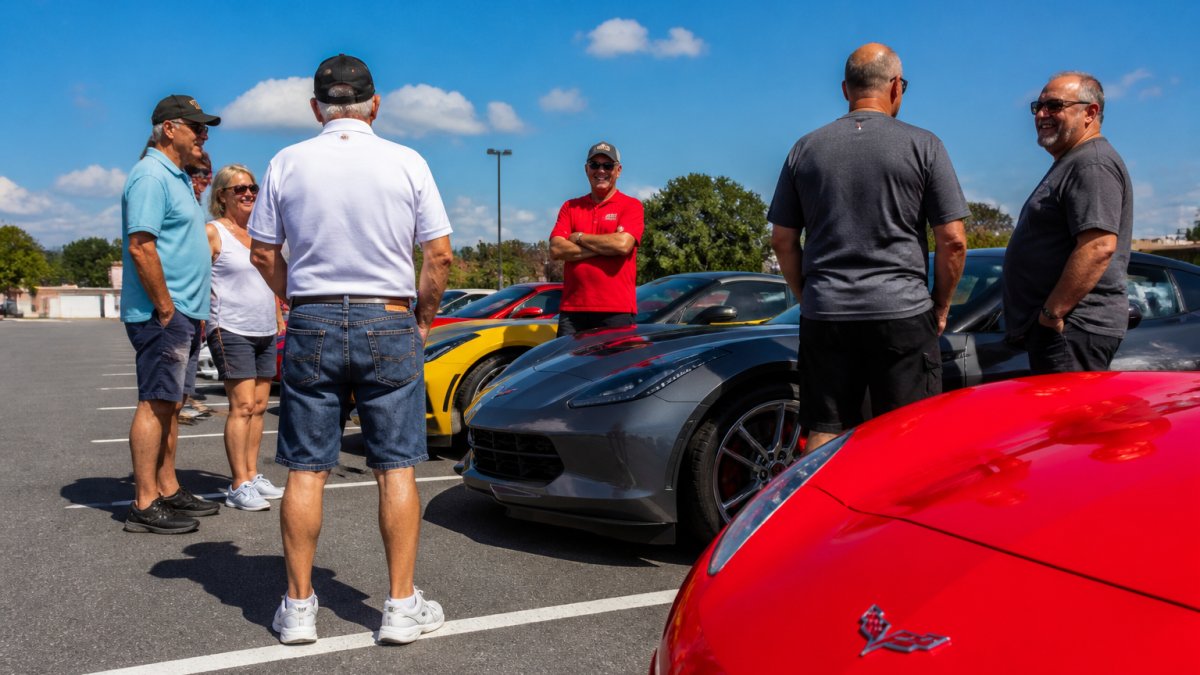 Group of Corvette owners chatting and smiling by their cars at a sunny Corvette clubs meetup in a parking lot