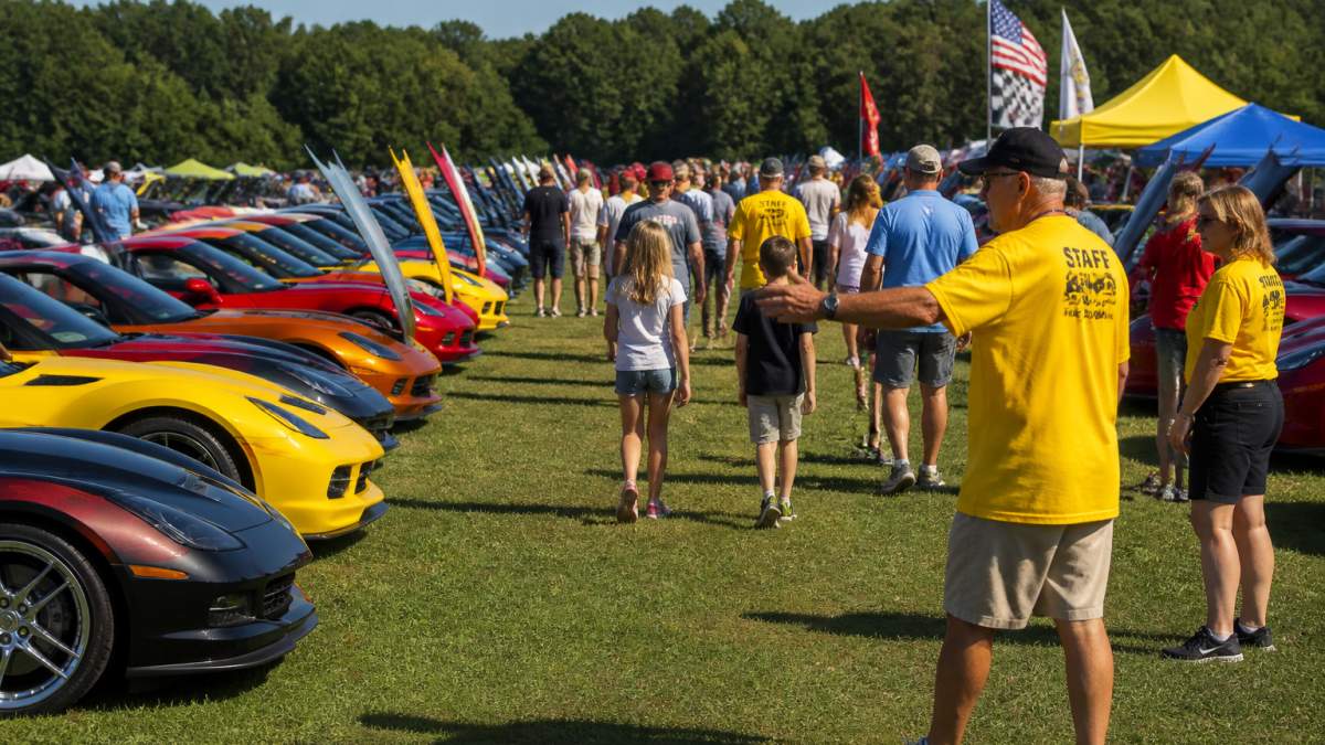 A line of colorful Corvettes parked on a sunny show field with volunteers in matching shirts directing attendees and families walking between the cars