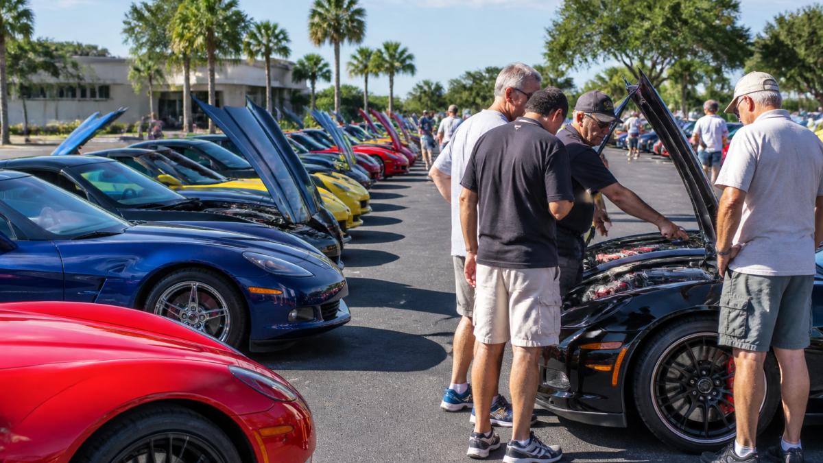 Owners discuss engine details beside colorful cars at Corvette meetups in a sunny parking lot.